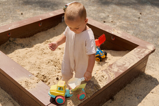 A child is playing in a sandbox at a park. The child is wearing a beige outfit and is using a toy truck to move sand. Trucks are nearby