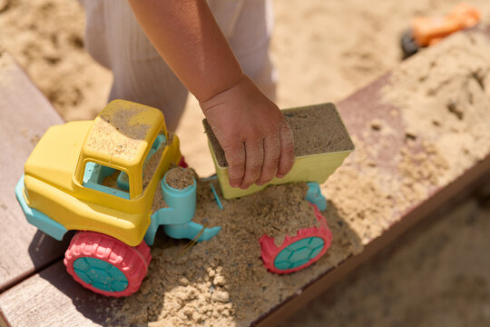 A young child is using a toy truck to move sand in a sandbox. The sun is shining and other toys are visible in the background