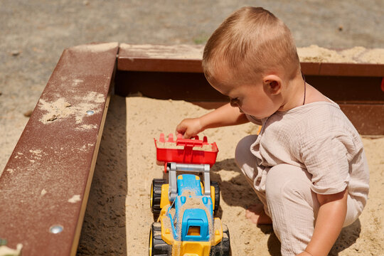 A young child sits in a sandbox digging and moving sand with a toy truck. The sun shines down on the playground, creating a warm atmosphere for play