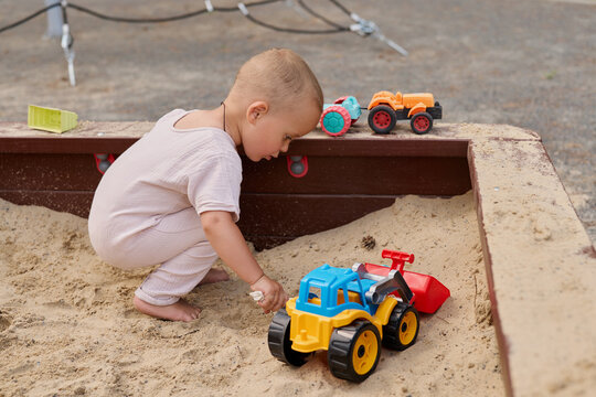A young child is playing in a sandbox at a park. The child is focused on moving toy trucks. Other toys are scattered around in the sand