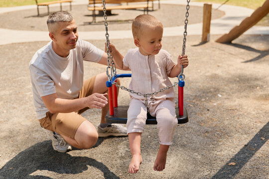 A child sits on a swing with bare feet while an adult stands nearby at a playground. The setting has gravel ground and other playground equipment