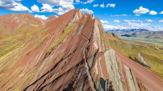 Aerial view of Vinicunca, the Rainbow Mountain, featuring vibrant mineral-rich sedimentary layers and rugged Andean terrain under a bright blue sky in Quispicanchi Province, Cusco, Peru.