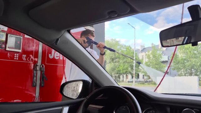 Man washing a car with a pressure hose at a self-service car wash, seen through the wet windshield from the driver's seat inside another vehicle.