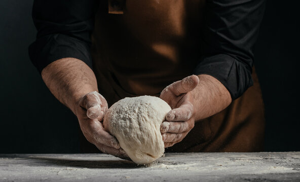 Male baker in brown apron holds kneaded dough with flour-covered hands over a rustic wooden countertop in a dimly lit kitchen setting