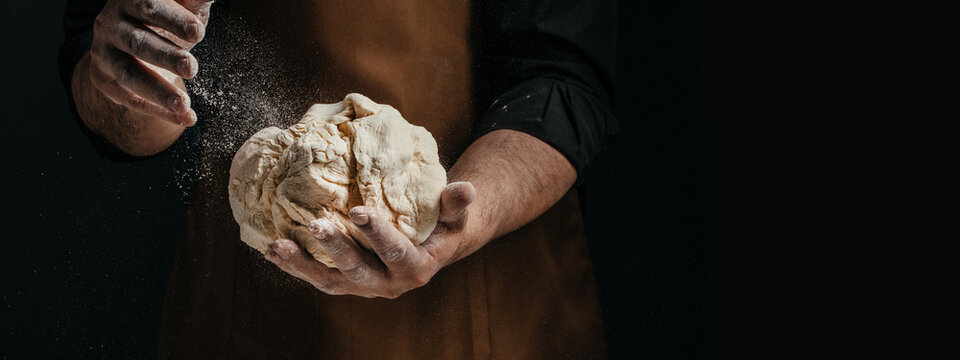 Male baker wearing brown apron holds kneaded dough with flour dusting in a dark kitchen setting, showcasing hands and texture of the dough