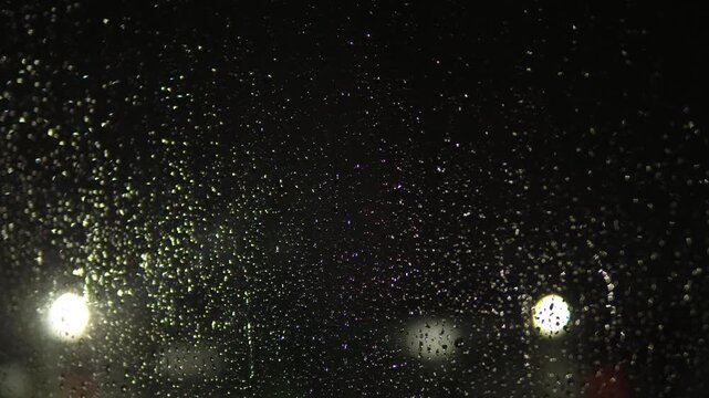 Numerous small raindrops cover the glass against a night street with bright blurred lights. Blurry lanterns and car headlights create a soft bokeh and a deep evening atmosphere