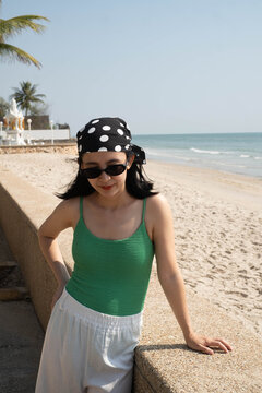 Young woman wearing sunglasses and a polka dot headscarf relaxing by a sandy beach on a sunny day, enjoying a peaceful ocean view. Summer vacation, coastal lifestyle, travel, wellness, and leisure.