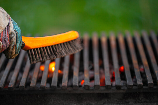 Hand wearing protective glove scrubbing a barbecue grill with a brush, glowing embers visible beneath the grates in an outdoor setting