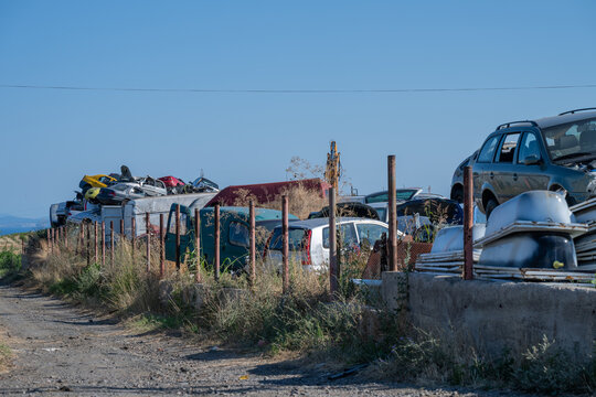 an overgrown auto salvage yard enclosed by a rusted metal fence, with various dismantled vehicles and scrap materials piled behind it along a dirt path.