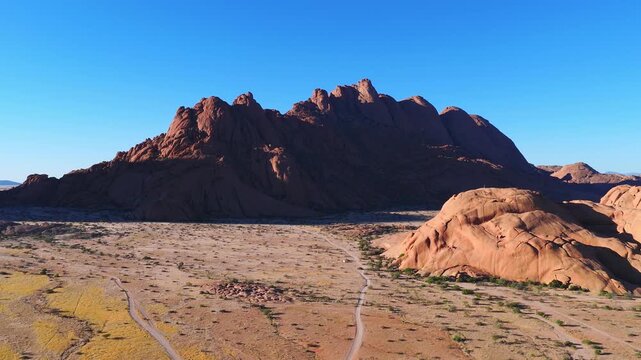 Aerial glide over a flat desert plain toward Spitzkoppe in Erongo, Namibia. Vehicle tracks and shrubs appear as long shadows fall, in warm light with wide framing.
