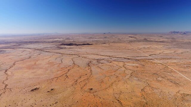 Aerial view of Spitzkoppe plain in Namibia shows rust orange gravel, braided dry riverbeds, inselbergs, and one pale dirt track, with slow sweeping motion in daylight.