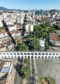 Aerial view of Arcos da Lapa with its iconic white arches spanning the urban landscape, surrounded by dense city buildings, lush green trees, and streets in Rio de Janeiro, Rio de Janeiro, Brazil.
