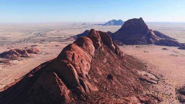 Aerial flyover of Spitzkoppe in Namibia shows reddish granite inselbergs, dome peaks, arid plain with shrubs and tracks, clear sky, crisp daylight, and steep shadows.