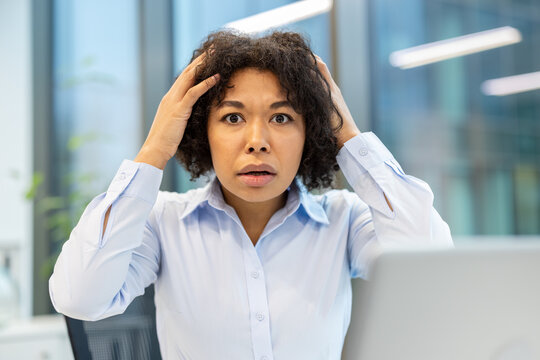 Portrait of a young, scared businesswoman sitting in the office, holding her head in her hands and looking at the camera
