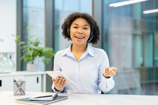 Smiling African American office worker sitting in the office in front of the camera wearing a headset, holding a tablet in her hands and talking
