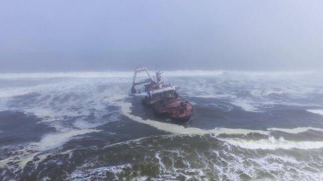 Aerial drone pulls back and pans around a rusted Zeila Shipwreck off Namibia's Skeleton Coast as heavy surf batters the corroded hull, fog softens a wide, high angle view.