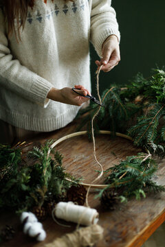 Female hands cutting thread with scissors to make Christmas wreath on rustic wooden table,close up. Florist workshop process, winter holiday decoration