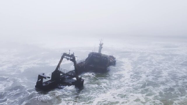Aerial view shows a rusting Zeila Shipwreck with A frame gantry and leaning mast on the Skeleton Coast, Namibia, as waves churn and fog drifts while the camera circles.