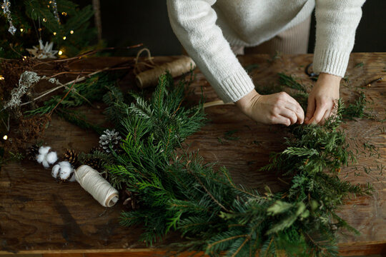 Woman making Christmas wreath with green fir branches and wooden hoop on rustic table. Cozy winter workshop, handcrafted xmas decor. Atmospheric holiday details