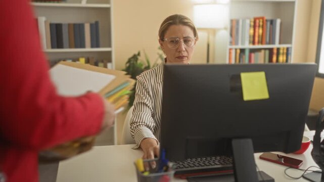 Woman at computer receiving a stack of folders from a colleague, hands reaching across a desk inside a building; workload stress.