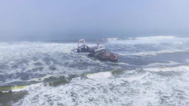 Aerial scene shows a rusting wreck with A frame crane listing in surf on Namibia Skeleton Coast as Atlantic waves surge and break in cool misty daylight tones.