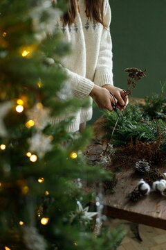 Making Christmas wreath. Woman cutting dried flowers with scissors for eco friendly xmas wreath. Cozy Christmas DIY workshop, warm bokeh xmas tree lights on background