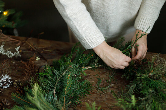 Making christmas rustic wreath. Female hands arranging fir branches to craft Christmas wreath on wooden table. Cozy DIY xmas workshop, natural eco friendly holiday home decoration.