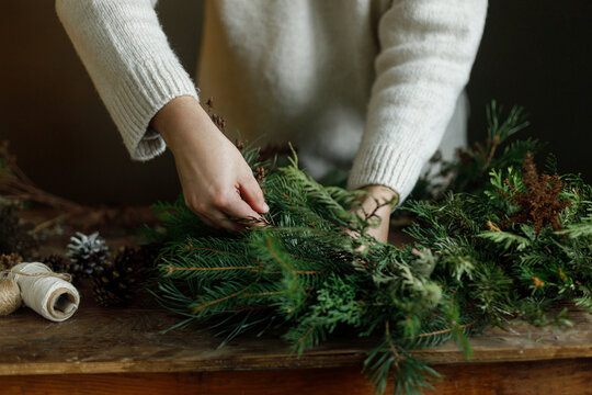 Making christmas rustic wreath. Female hands arranging fir branches to craft Christmas wreath on wooden table. Cozy DIY xmas workshop, natural eco friendly holiday home decoration.