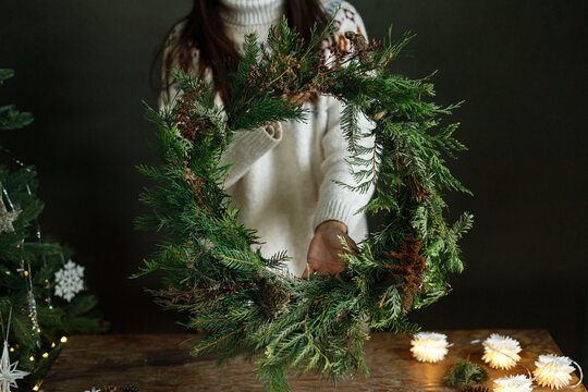 Woman in cozy sweater holding finished handmade Christmas wreath near decorated xmas tree with warm lights. Festive DIY workshop, winter holiday home decoration