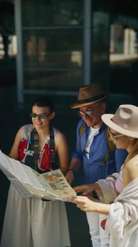 Happy tourists consult a map, wearing hats and sunglasses outdoors. Friends are exploring their travel destination and planning their adventure.