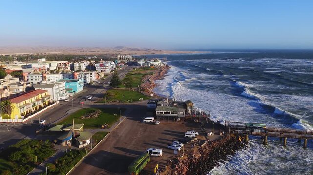 Aerial view of Swakopmund, Namibia, with the iconic jetty, surf on a rocky seawall, a pier restaurant, pastel colonial buildings, a promenade, dunes, and a seaside park.
