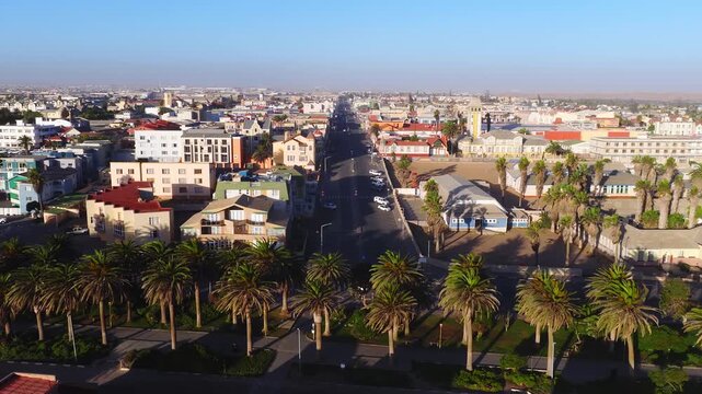 Aerial view of Swakopmund, Namibia, as a drone glides along a palm lined seafront road, pastel houses, colonial buildings, a yellow tower, and parked white cars appear.