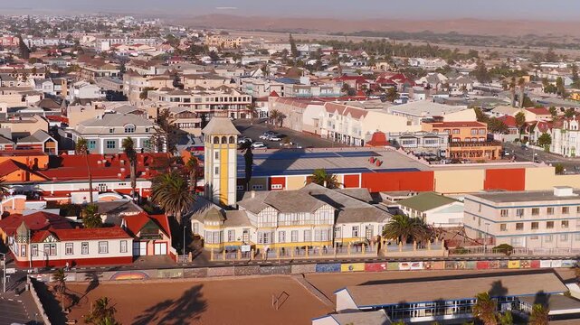 Aerial view of Swakopmund, Namibia, with German era architecture, yellow and white tower, murals on the seawall, palm lined beach, and Namib Desert dunes in warm light.