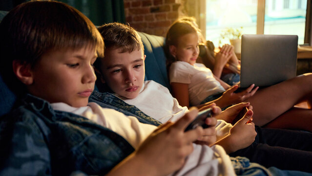 Boys using smartphones while another child works on laptop in background. Concept of mixed-device leisure, parallel online activity, connected childhood.