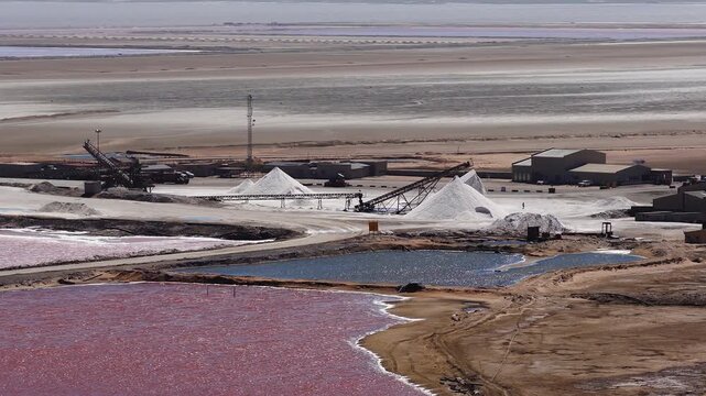 Aerial view of Walvis Bay salt pans in Namibia shows pink and blue ponds, tidal flats, conical white salt piles, warehouses, and conveyors in bright midday light.