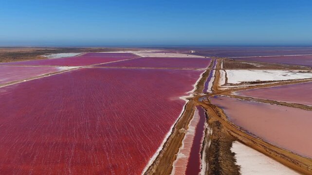 Aerial view of Walvis Bay salt works in Namibia shows red and pink pans, earthen levees, white salt beds, service tracks, salt piles, ocean horizon, and rippling brine.