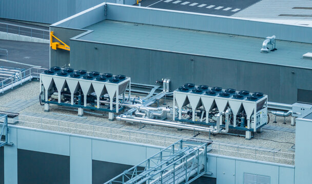 Aerial view of industrial air-cooled chillers with multiple axial fans and insulated piping installed on the rooftop of a modern manufacturing facility in Grenoble, Auvergne-Rhone-Alpes, France.