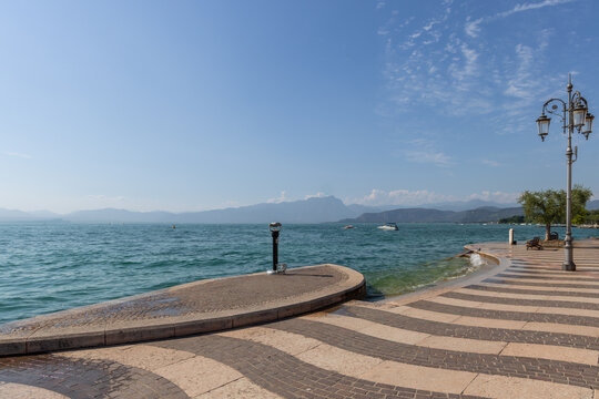 Wave-pattern paving and ornate lamps line Lungolago Marconi in Lazise, Italy, as the promenade curves along Lake Garda toward open mountain views.