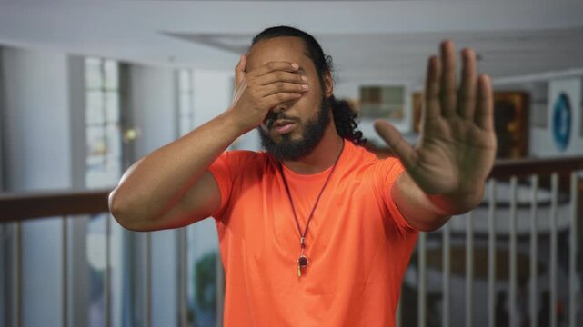 Man in an orange shirt covering his eyes with one hand and holding the other palm out in a hotel building corridor, hands raised in stop gesture; privacy refusal.