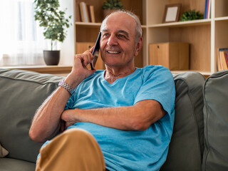 Fototapeta premium Portrait of happy senior man smiling and enjoying talking on the mobile phone while relaxing on sofa at his cozy home.