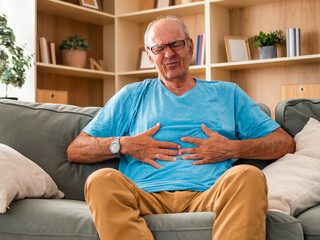 Fototapeta premium Senior man sitting on sofa in his living room and holding his abdomen with a painful expression. He is illustrating stomachache, indigestion, digestive problems and discomfort at home.