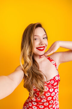 Smiling young woman taking a selfie in a red daisy dress against a bright yellow background, playful summer studio portrait