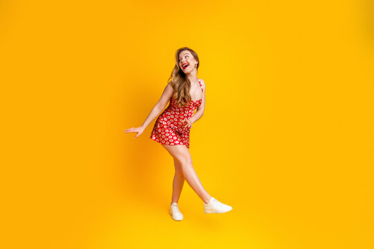 Young woman in red floral summer dress smiling and posing against a bright yellow studio background, playful fashion portrait with white sneakers and confident cheerful expression
