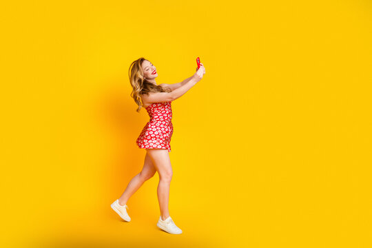 Young woman in red floral dress taking a cheerful selfie against a bright yellow background, playful summer portrait with casual sneakers and vibrant color