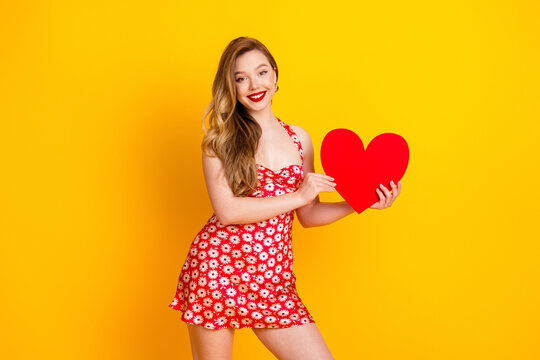 Smiling young woman in red floral dress holding a large red heart against a bright yellow background for Valentine and romantic lifestyle concept