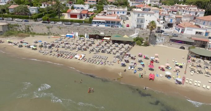 Aerial view of a sandy beach resort lined with rows of sun umbrellas, sunbeds, beach clubs and people enjoying the calm sea waters on a bright summer day. It's San Felice Circeo beach, in Lazio, Italy