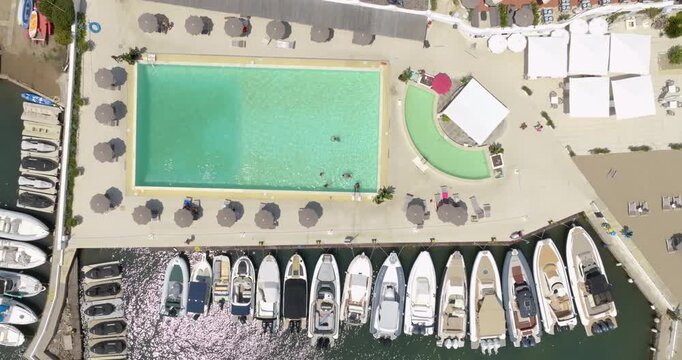 Aerial top-down view of a luxury coastal resort featuring a rectangular swimming pool surrounded by sun umbrellas and lounge areas, situated right next to a marina with docked boats and yachts.