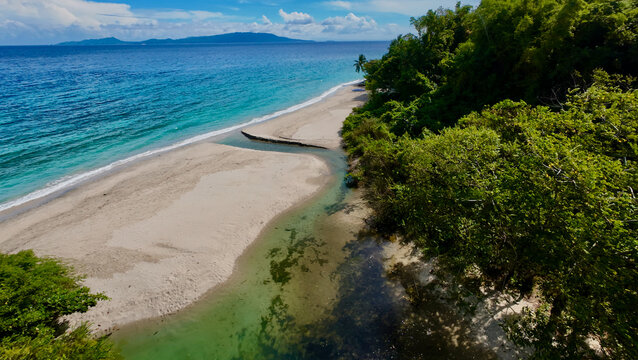 A tropical beach meets a winding stream on a quiet coastline. Aerial view shows clear turquoise water, white sand and lush forest where a small river flows into the sea.