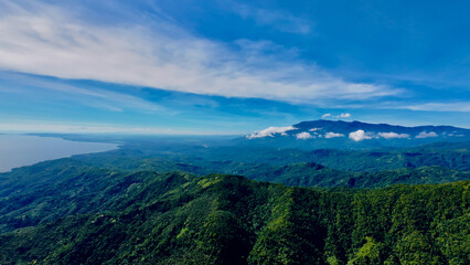 Fototapeta premium Mountain ranges stretch to the distant coast beneath a wide blue sky. Layers of green hills and drifting clouds create a dramatic tropical aerial landscape.