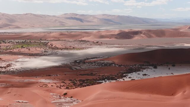 Aerial drone glide over Sossusvlei and Dead Vlei in Namibia shows red dunes, pale salt pans, distant mountains, tiny hikers on a crest, and soft daylight with clouds.